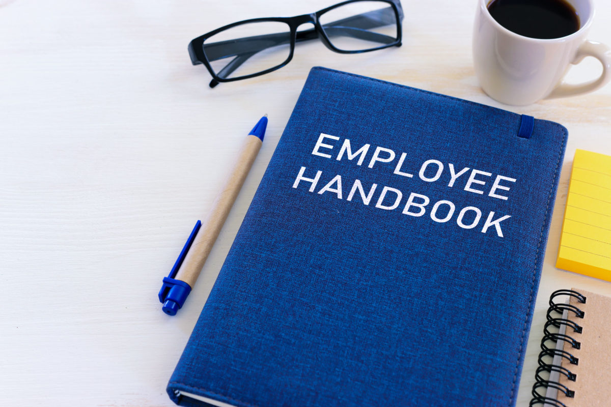 Employee handbook on desk next to sticky notes, pen, reading glasses, and coffee